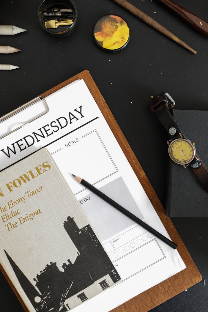 Flat lay of a workspace with book, planner, pencil, and wristwatch on a wooden surface.